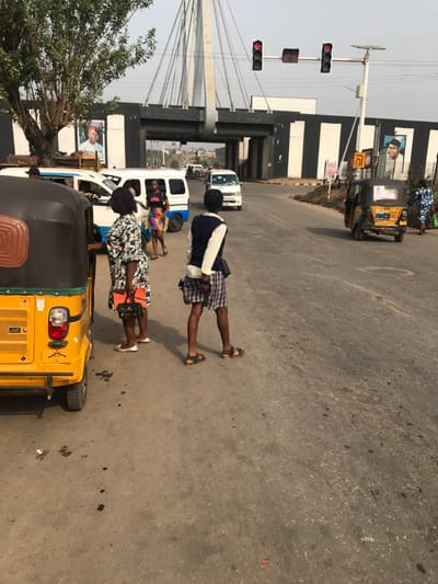 The image presents a daytime street scene at an active intersection in Umuokpu, Nigeria.
In the foreground left, a yellow and black three-wheeled auto-rickshaw, branded "AXIS" with a partially visible license plate, is stationary on the asphalt roadway. Adjacent to it, a female subject in a patterned dress, wearing light-colored shoes and holding an orange and black handbag, is positioned with her side to the camera, appearing to interact with the vehicle. To her right, a second subject, dressed in a white long-sleeved shirt under a dark vest, plaid shorts, and dark sandals, stands on the roadway with their back to the camera, facing right. The asphalt road surface displays visible wear, minor cracks, and dark stains.
In the mid-ground, a white and blue passenger minivan is visible on the left, with multiple occupants. A female subject in a dark top with colorful patterns and a colorful skirt is standing near this minivan. Another white minivan is further back in the center. On the right side of the road, a second yellow three-wheeled auto-rickshaw, also branded "AXIS" (license plate SY2 131), is present. Further right, a female subject in a blue patterned dress walks away from the camera on the road shoulder.
The background is dominated by a prominent cable-stayed overhead bridge, featuring white and gray structural elements and black and white striped support pillars. Above the main road, three traffic lights are visible, two displaying red circles and one displaying a red left-turn arrow. These lights are powered by a solar panel array mounted on a pole to the right. Beneath the bridge structure, large billboards featuring a smiling woman with a red headwrap (left) and a man in a white cap (right) are affixed to the support walls. A large tree with green foliage stands on the left, partially obscuring the left billboard. Distant urban architecture and undulating terrain are discernible beneath the bridge structure.