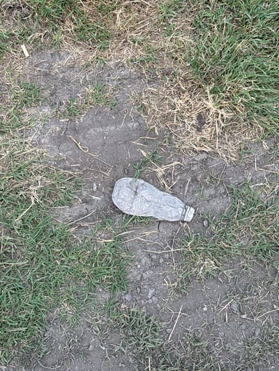 A discarded, flattened plastic water bottle lies on a patch of dry, cracked earth surrounded by sparse, green grass. The bottle is translucent and appears to be empty. The ground is a mix of soil and dry grass, with some small pebbles visible. A single cigarette butt is also present to the left of the bottle. The overall scene suggests an outdoor environment, possibly a roadside or park area, in Fontevivo, Italy.