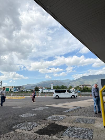 This image depicts an outdoor scene, likely a rest stop or service area in Castrocielo, Italy, under a bright, partly cloudy sky. The perspective is slightly angled, showing the underside of a dark gray building overhang in the upper left.
In the foreground on the right, a man in a blue and white striped shirt and jeans stands next to a yellow metal railing. The ground is paved with a combination of asphalt and cobblestones, featuring several metal manhole covers. One prominent manhole cover clearly reads "ITALIANA GAS ACQUA GENOVA."
Towards the center of the image, a white commercial van is parked. To the left of the van, two individuals are visible further back: one wearing a blue top and the other in a floral skirt with a purple top. On the far left, a man in a plaid shirt and jeans is leaning against a low barrier or structure.
The background is dominated by large, green mountains or hills under a blue sky with scattered white clouds. Several buildings, including what appears to be a service station or industrial structures, are visible in the mid-ground. Notable signage includes a green sign with "AUTOGRILL" and a blue banner that reads "BLU TRASPORTI." A tall streetlight pole stands centrally. The overall impression is a daytime scene with no specific activity beyond people resting or waiting in an open, functional area.