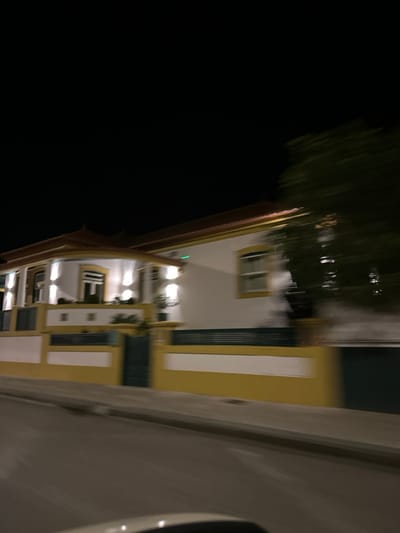 A building at night features multiple lit windows and white and green accents against a yellow facade. Dark skies surround the structure, with blurred vegetation possibly reflected in a nearby body of water. A section of a white wall, possibly a structure or vehicle, is visible on the right side of the image.