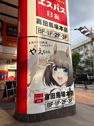 This image depicts a street-level view of a cylindrical pillar advertising the pachinko and slot parlor "Espass Takadanobaba Main Store" in Shinjuku, Japan. The pillar is covered with large, colorful advertisements. Prominently featured is an anime-style illustration of a young woman with brown hair, large blue eyes, and horse ears, giving a peace sign. The text on the pillar includes "Espass" in large red lettering, along with the store's name "高田馬場本店" (Takadanobaba Honten) and floor information "BF・1F・2F・3F". Other advertisements on the pillar include "快適CLUB" (Kaiteki CLUB) and "コミック＆ネット 鍵付完全個室" (Comic & Net Keyed Private Rooms) with "24h OPEN".

In the background, a modern building with multiple windows and air conditioning units is visible. A street lamp with multiple light fixtures is attached to a pole on the right side of the image. A black van is parked on the street. The ground is paved with cobblestones and concrete. The overall scene is set outdoors, suggesting a commercial district. The lighting indicates it is daytime, with some shadows present, possibly late afternoon.