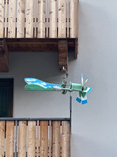 A miniature windmill decoration is mounted on a pole outside a building, likely a residential property, in La Ila - Stern - La Villa, Italy. The windmill is designed to resemble a person piloting a stylized airplane. The "plane" element is a long, narrow piece painted to depict a green landscape with a blue sky and white clouds. A small figure, dressed in a hat and green clothing, stands at the front of the "plane" as if steering it. The windmill's rotating blades are painted with alternating green and blue and white sections. The entire structure is positioned against a light gray textured wall, with a wooden balcony railing in the foreground and another wooden structure above, suggesting it's on a balcony or exterior wall of a multi-story building. A dark green door is visible in the background to the left.