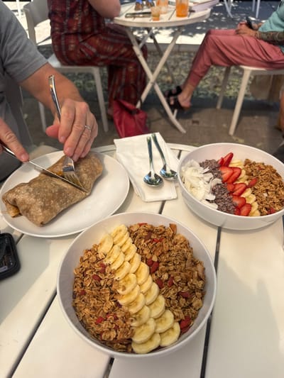 The image depicts an outdoor dining scene in the city of Bend, United States, viewed from a slightly elevated perspective. In the foreground, a person wearing a grey shirt and a wedding band on their left ring finger is actively cutting a large burrito on a white plate with a knife and fork. To the right of the burrito, on a white slatted table, are two white bowls containing acai or smoothie bowls. The bowl closer to the viewer features granola, goji berries, and neatly fanned slices of banana. The second bowl contains granola, sliced strawberries, sliced bananas, dark cacao nibs or chocolate chips, and large white coconut flakes. A folded white napkin holds two small silver spoons between the burrito plate and the bowls. A dark smartphone is partially visible on the table to the far left.
In the background, other patrons are seated at white outdoor tables. One person to the left wears red patterned trousers, with a red bag positioned on the ground near their feet. Another person to the right wears pink trousers and has a visible tattoo on their right arm. On the white round table in the background, two glasses containing orange-colored beverages are visible. The ground beneath the tables appears to be paved with dark pavers or cobblestones, and shadows suggest daytime lighting.