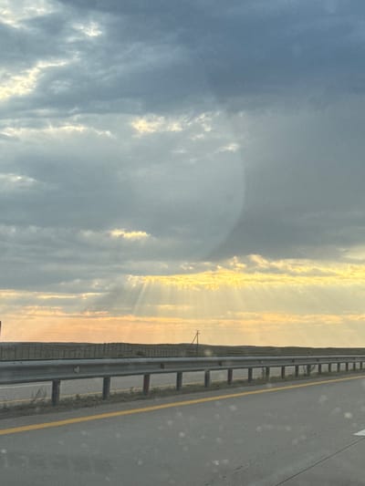 The image depicts a highway scene at dusk or dawn, with the sun's rays breaking through a cloudy sky. The sky is filled with a mix of dark and light gray clouds, with streaks of golden light visible as the sun shines through. The sun is low on the horizon, casting a warm glow over the scene.
In the foreground, there is a paved highway with a yellow line marking the lane. A metal guardrail runs along the side of the road, supported by vertical posts. Beyond the guardrail, there is a flat, open landscape with sparse vegetation. A fence or barrier is visible in the distance. The overall impression is one of a rural or semi-rural highway. The specific location in Торғай стансасы, Kazakhstan cannot be determined from the image alone, as the visual elements are not unique to any particular city.