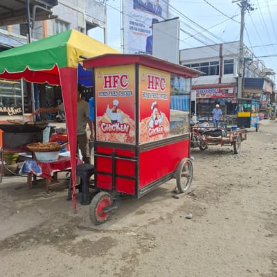 In Rhieng Blang, Indonesia, a street vendor operates a food cart selling fried chicken under a green and yellow awning. The cart itself is red and yellow, featuring prominent branding for "HFC Husen Fried Chicken" with animated chicken graphics. In the background, several buildings line the street, including shops displaying "ACCESSORIES" and "IMITASI DAN MAINAN." Power lines crisscross overhead, indicating an urban or semi-urban environment. A man in a blue shirt and cap walks past a motorcycle and trailer, suggesting daily commerce. The ground is dirt and gravel, typical of unpaved roads. Another vendor is seen cooking at a wok, surrounded by cooking implements and food preparation materials. The scene captures a moment of everyday life and commerce in the specified Indonesian locale.
