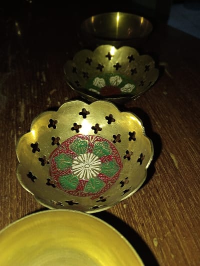 The scene, set in Longmiang, Laos, depicts four small brass bowls arranged on a dark wooden surface. In the mid-ground, a prominent brass bowl features a scalloped rim with perforated cross-shaped cut-outs. Its interior is adorned with an intricate, enamel-like design centered around a white chrysanthemum-like flower, encircled by green leaves, all set against a red patterned background. Immediately behind this, a second, similarly styled scalloped brass bowl is partially visible, appearing slightly out of focus. In the lower foreground, the smooth, curved edge of a plain, unadorned brass bowl is visible, partially cropped from the frame. Further in the background, a fourth, simple brass bowl is faintly discernible and largely obscured. The wooden surface exhibits visible grain and minor imperfections. The environment is dimly lit, with reflections of light visible on the polished brass surfaces.