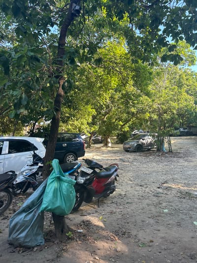 The image depicts an outdoor parking area in A R A, Indonesia, beneath a canopy of leafy trees. In the foreground, several motorcycles are parked, with one white motorcycle bearing a "SUZUKI" logo visible. Two large bags, one light blue and one green, are tied to the trunk of a tree on the left side of the frame. Behind the motorcycles, a white car and a dark blue SUV are parked. Further into the background, a gray car is visible, along with other vehicles partially obscured by trees and foliage. The ground is a mixture of dirt and gravel, with scattered leaves. The overall scene suggests a moderately sunny day, with dappled sunlight filtering through the trees. There are no people visible in the image.