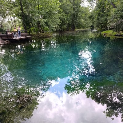 This is a photograph of a serene natural scene with a striking, crystal-clear turquoise lake surrounded by lush green trees. A wooden platform juts out over the water on the left side of the frame, where four individuals are standing. One person in a yellow shirt is taking a photo with their phone, while another in a blue shirt and white pants stands nearby. Two other individuals, one in a green shirt and another in a dark top, are also on the platform, appearing to be observing the water. The water itself is exceptionally transparent, revealing the lakebed below and reflecting the vibrant green foliage of the trees and the white, fluffy clouds in the sky. The reflections are so clear they create a mirror image of the surroundings, almost indistinguishable from the actual scene above the waterline. The overall atmosphere is peaceful and tranquil, suggesting a pleasant day in a park or natural reserve, possibly in the late morning or early afternoon given the bright, diffused light. The location is identified as Sdt Shcherbakovka, Russia. There is no visible text in the image.