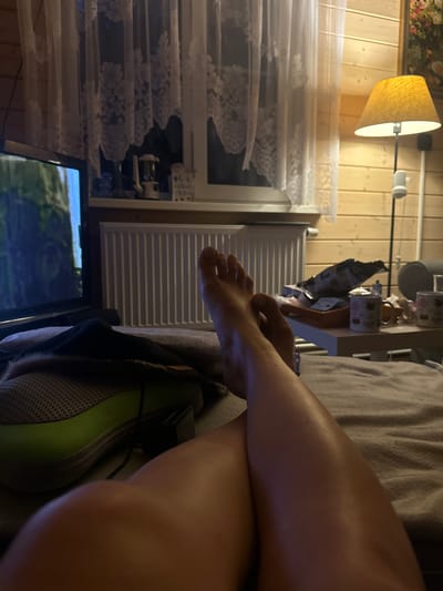The image depicts a dimly lit indoor scene, likely a living room or bedroom, with wooden paneled walls. In the foreground, a person's legs and feet are crossed, resting on a bed or couch covered with a gray blanket. A television screen is visible on the left, showing a nature-related image. To the right, a side table holds several mugs, a tray with items, and a lamp with a yellow shade. A white radiator is positioned below a window with lace curtains. The overall impression is one of relaxation and leisure, with the television and comfortable seating suggesting a home environment. There is no discernible connection to the city of Stupino, Russia.