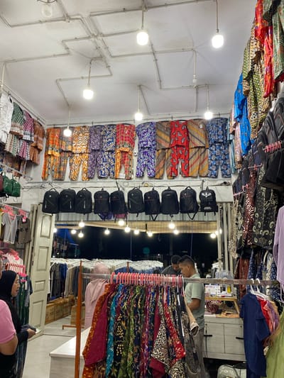 This media file shows an indoor market stall filled with various textiles and bags. The subjects include at least three people. One person, a man wearing a light blue polo shirt and dark pants, is examining textiles. Another person, a woman wearing a black hijab and a pink top, is looking at her phone while sitting. A third person, also wearing a black hijab, is partially visible in the foreground. The stall is brightly lit by overhead lights, and the textiles are displayed on racks, featuring colorful patterns, likely batik, common in Indonesia. Bags are also stacked on shelves and hung on walls.
The scene appears to be a retail environment, possibly a market or a shop. The setup suggests a commercial activity, with the people potentially being a vendor and customers. The overall atmosphere is that of a busy marketplace, though the exact time of day is not clearly discernible. However, the dark background visible through an opening suggests it might be evening or night. There are no overt weather cues.
Notable details include the abundance of intricately patterned fabrics, the various types of bags, and the electrical conduits running along the white ceiling. Some boxes with visible text, potentially shipping boxes, are stacked in the background. Although the text on the boxes is not entirely legible, one might contain the word "SOU". The interaction of the people with the goods implies shopping or selling activities are underway. The image conveys a sense of commerce and local culture, given the prominent display of batik-style fabrics. The location context of Lamteumen Barat, Indonesia, further reinforces the likely presence of such textile markets.