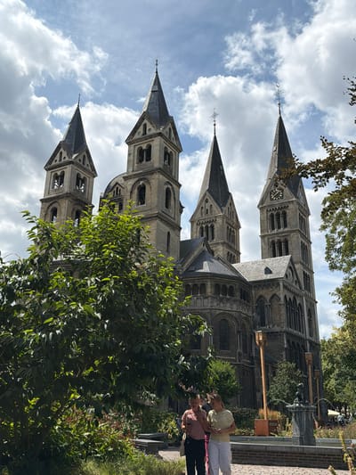 In Roermond, Netherlands, a large stone church with multiple spires dominates the background, under a cloudy sky. A lush green tree is in the foreground, obscuring part of the church's lower section. Two individuals are standing in front of the tree, appearing to be engaged in conversation. A fountain is visible to the right of the people, and foliage lines the area around it. The architecture suggests a historical European city.