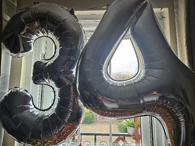 The image displays two large, silver, reflective foil balloons, inflated and forming the number "34." These balloons are positioned indoors, in front of a window, which serves as the primary light source and reveals an outdoor scene. The indoor environment includes dark beige curtains visible on both sides of the window. Through the clear windowpanes, green foliage, possibly trees or shrubs, is visible in the background under an overcast sky. A black metal railing or fence is present directly outside the window at a lower level. On a ledge or surface beyond the railing, a white plant pot containing pink flowering plants, consistent with geraniums, is visible. The reflective surfaces of the balloons show distorted reflections of the surrounding indoor space and the outdoor view. No people are visible in the scene. The specific city within Cherwell District, United Kingdom, cannot be determined from the visual evidence.