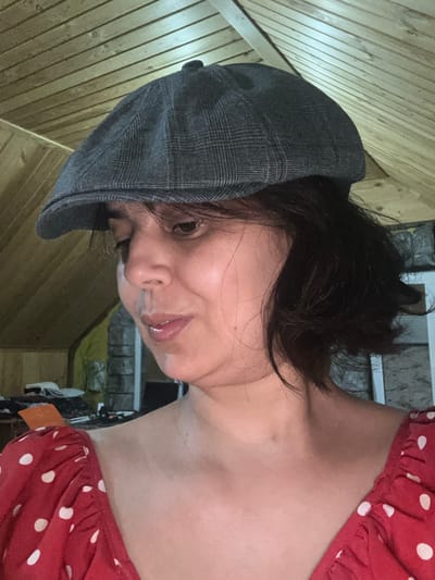 A medium shot captures a woman wearing a plaid flat cap and a red polka dot dress. She is looking down and to her left, with her mouth slightly open. Her dark hair is cut short and appears to be slightly tousled. The background features a wooden ceiling and a stone wall. The scene is indoors, possibly in an attic or loft space. There are no discernible elements in the image that would indicate the location within Kalininskaya, Russia.