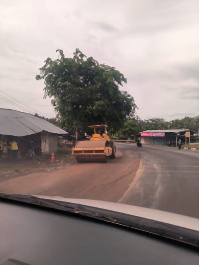 The image captures a daytime scene in Seulimeum, Indonesia, from the perspective of a car's dashboard, showing a road under construction or repair. The sky is overcast, indicating cloudy weather.

The primary subject is a large, yellow road roller (compactor) prominently positioned on the unpaved left side of the road, facing away from the viewer. The brand "SAKAI" is visible on its side. A person can be seen operating the roller from the driver's seat.

To the left of the road roller, several people are gathered near modest buildings with corrugated metal roofs. One individual appears to be wearing a yellow vest. An orange traffic cone is placed on the dusty ground nearby. The immediate foreground shows the vehicle's dashboard and windshield wipers.

The road ahead curves to the right, transitioning from the unpaved section to a paved surface. Further down the paved road, a person on a motorcycle is driving away from the viewer. A black car is parked near another building on the right side of the road, where a person is also standing. This building displays a banner with the text "RAMADAN BERKAH." Lush green trees dominate the background, characteristic of a tropical environment. Faint power lines are visible in the upper left. The scene depicts ongoing infrastructure work integrated with daily activities in a semi-rural setting.