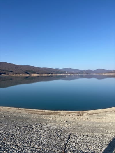 The image, correctly oriented, depicts a serene landscape featuring a large body of water, distant mountains, a clear blue sky, and a gravelly bank in the foreground.

The upper portion of the frame is dominated by a vast, cloudless blue sky, indicating bright and clear weather conditions. This deep blue transitions into a lighter hue closer to the horizon.

Across the water, a range of mountains or hills forms the distant shoreline. These landforms appear rugged and are mirrored almost perfectly in the exceptionally still, dark blue surface of the water. The reflections are so clear that they create an elongated, symmetrical visual effect, blending the natural landscape with its aqueous counterpart.

The middle section of the image is filled by the calm, deep blue water, which exhibits a smooth, almost glassy surface. Its stillness allows for the crisp reflections of the sky and mountains.

In the immediate foreground, occupying the lower right side of the frame, is a textured bank or dam wall. This area is covered in light-colored gravel and small stones, which are illuminated by direct sunlight, casting subtle shadows that highlight their rough texture. The bank appears to gently slope towards the water.

There are no people or specific activities visible in the image, emphasizing the natural tranquility of the scene. The time of day is clearly daytime, characterized by strong, natural illumination. No visible text is present. Given the provided context, the scene is likely a view of the Sioni Reservoir in Sioni, Georgia.