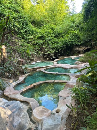 The image shows a series of terraced hot spring pools nestled within a lush, green environment. The pools are constructed with natural-looking stone and concrete borders, with clear, turquoise water. Vegetation, including trees and dense foliage, surrounds the pools, suggesting a natural or semi-natural setting. The lighting indicates it is daytime, with sunlight filtering through the leaves. The scene appears to be a tranquil and scenic location.