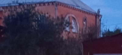 An exterior, medium shot captures a building in Novokabanovo, Russia, under a twilight sky. The building, constructed of brick, features a red facade and a grey, sloped roof. Distinctive architectural elements include a row of decorative triangular protrusions along the upper edge of the building, and two arched windows. One window, larger and more centrally located, has a pattern of lighter and darker shades within its arch. To its right, a smaller, narrower arched window is visible. The building is partially obscured by dark foliage in the foreground. To the right of the main building, a portion of another, darker red structure with a peaked roof is visible. The scene is dimly lit, suggesting either dusk or dawn.