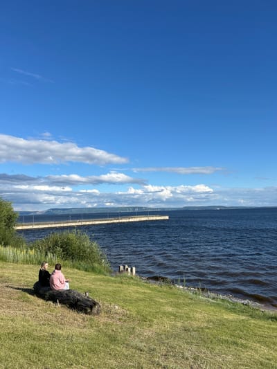 The image shows two people sitting on a log on a grassy embankment overlooking a body of water. They are facing away from the camera. The water is dark and appears to have small waves. In the distance, a long pier extends into the water, with lights visible along its length. Beyond the pier and water, there is a distant shoreline with hills. The sky is blue with scattered white clouds. The scene appears to be outdoors on a clear day. The location is identified as Primorsky, Russia.