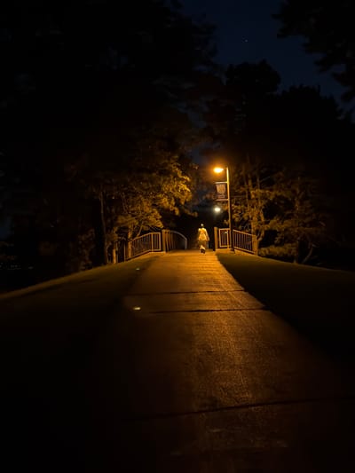 A person walks alone on a wet bridge at night, illuminated by a single lamp post. The bridge has wooden railings and a paved walkway. Trees surround the bridge, their dark silhouettes stark against the night sky. A street lamp with a banner attached casts a warm yellow light on the bridge, creating reflections on the wet surface. The sky is dark with a few visible stars. The person, dressed in light-colored clothing, walks away from the camera towards the far end of the bridge. The scene suggests a quiet, possibly peaceful, atmosphere. The location is identified as the village of Vernon Hills, United States of America.
