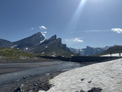 This image depicts a mountain landscape near Leukerbad, Switzerland. A river flows through the foreground, with rocky banks and patches of melting snow. A stone dam or wall crosses the river in the middle ground, with two hikers walking on top of it. Beyond the dam, more mountains rise, some with snow-capped peaks. The sky is clear and blue with some scattered clouds, and the sun is shining brightly, creating lens flare in the upper right corner. The scene appears to be captured on a sunny day.