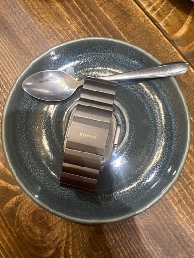 The image shows a grey-blue glazed ceramic bowl resting on a wooden surface. Inside the bowl, there is a silver spoon and a smartwatch with a dark grey metal link band. The band is branded with the word "NOMAD". The spoon lies across the bowl, partially on top of the watch band. The lighting is bright, creating reflections on the spoon and the bowl's surface. The wooden surface appears to be a table with a light brown and dark brown wood grain pattern.
The image does not contain information to determine its location within Mole Valley, United Kingdom.