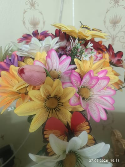 A close-up, vertical shot showcases a vibrant artificial flower arrangement. The bouquet is a colorful medley of daisies and a single pink tulip. The daisies display a variety of hues, including bright yellow with detailed brown centers, pink with white edges and intricate veining, orange, and white. Small white flowers with green stems are interspersed throughout. Two artificial butterflies, one predominantly red with orange and black accents, and another with yellow and black patterns, are positioned among the flowers, adding a whimsical touch.

The backdrop is a light beige wall with a subtle, ornate damask pattern, suggesting an indoor setting, possibly a home or an event space. The lighting appears to be artificial, casting a soft glow on the arrangement. The lower right corner of the image contains visible text: "27.06.2026 19:51," indicating the date and time the photo was taken. The overall scene evokes a cheerful and decorative atmosphere, likely meant to enhance an interior space. The image is sharp, focusing on the details of the artificial flowers and butterflies. No people are visible in the frame. The location context is Qaskeleñ, Kazakhstan.