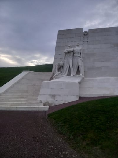 A monument of white stone stands prominently against a cloudy sky. The structure incorporates a sculpture of two figures draped in robes, positioned on a platform near the top of the monument. Steps lead up to the monument alongside a path with dark gravel. Lush green grass surrounds the monument on two sides.