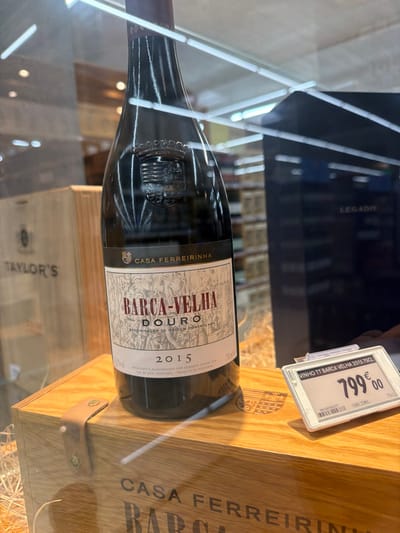 A dark glass bottle of "Barca Velha Douro 2015" red wine is presented within a retail display case in Lamego, Portugal. The bottle prominently features a label indicating "CASA FERREIRINHA" at the top, an etched-style illustration, and "BARCA-VELHA DOURO Denominação de Origem Controlada 2015 75cl," with production details for Sogrape Vinhos S.A. in Vila Nova de Gaia, Portugal. The bottle rests on a light-colored wooden presentation box, partially inscribed with "CASA FERREIRINHA BARCA" and secured with a leather strap and brass rivets. Adjacent to the wine bottle, a digital price tag displays "VINHO TT BARCA VELHA 2015 75CL" priced at 799.00 Euros. Shredded straw-like material is visible around the base of the bottle. The background, viewed through the glass, includes reflections of overhead fluorescent lighting, additional shelving stocked with bottles, a wooden box marked "TAYLOR'S" on the left, and a dark display unit with "LEGADO" visible on the right, indicating a high-end wine retail environment. No individuals are present.