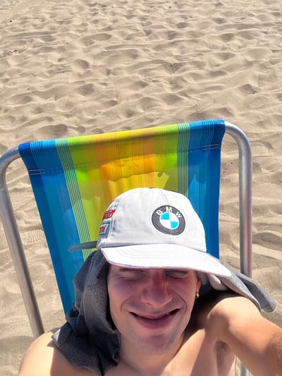 A man lies on a blue, yellow, and green striped beach chair with a light gray BMW baseball cap. The chair sits on a sandy beach. The man's face is visible, with eyes closed and a slight smile. He wears a dark gray shirt draped over his shoulder. The beach is light brown, with patterns of light and shadow from the sun.