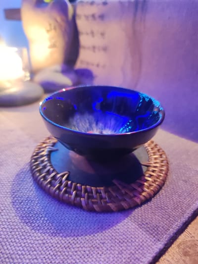 This image, captured in the city of Mas, Indonesia, displays a still life under dim, mixed lighting conditions. The primary subject is a dark, possibly navy blue or black, ceramic cup with a glossy interior, centrally positioned. The interior surface of the cup exhibits vivid blue and white light reflections, suggesting a highly reflective glaze or a complex internal pattern interacting with light. The cup rests on a circular coaster constructed from woven, light brown material, likely rattan. This coaster is placed on a light-colored, coarsely woven fabric mat or placemat, which in turn covers an indistinct, darker surface.
The scene is illuminated by multiple light sources. A dominant cool, purplish-blue light casts a glow over the foreground, the mat, and the exterior of the cup, contributing to the strong blue reflections inside the cup. In the blurred background, to the left, a distinct warm, yellow-orange light source, possibly a lamp or candle, emits a soft glow. To the right background, an indistinct, lighter vertical surface is visible, bearing faint, illegible markings or text. No individuals are present, and no actions or interactions are depicted. The environment is consistent with an indoor setting, possibly a restaurant, cafe, or private dwelling.