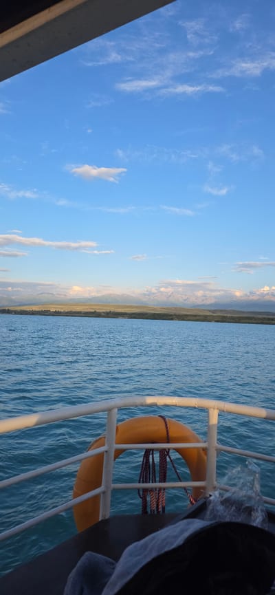 This media file captures a scenic view from a boat on a body of water. The sky is a vibrant blue with scattered wispy white clouds, suggesting a clear day. In the distance, a long stretch of land is visible, characterized by rolling hills and mountains that appear to be illuminated by the soft light of either sunrise or sunset. The water is a deep blue, with gentle ripples indicating calm conditions.
On the right side of the frame, a portion of the boat is visible, featuring a white metal railing. Attached to this railing is a bright orange life preserver, secured with a purple and red rope. A dark object, possibly a bag or clothing, is draped over the railing near the life preserver. The scene conveys a sense of tranquility and natural beauty. The time of day appears to be either early morning or late afternoon due to the soft lighting on the distant mountains. The location is identified as Pristan'-Przheval'sk, Kyrgyzstan, which is a port city on the eastern shore of Lake Issyk-Kul. There is no visible text in the image.