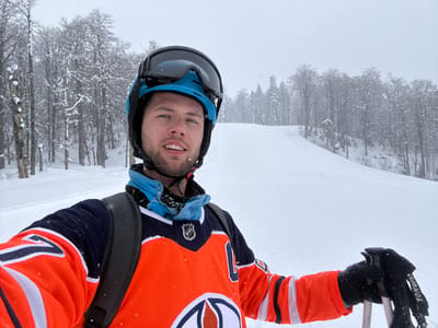 A young adult male is depicted in a self-portrait on a snow-covered ski slope, presumably in the vicinity of Mušovića Rijeka, Montenegro. He is wearing a light blue ski helmet with dark ski goggles pushed up, and an orange, navy blue, and white NHL Edmonton Oilers hockey jersey. A black backpack strap crosses his right shoulder. His left hand, covered by a black glove, holds two ski poles, one with a "ROSSIGNOL" brand strap visible. The man has a light beard and is smiling directly at the camera. The background features a wide, snow-covered ski run extending uphill, bordered by a dense forest of snow-laden deciduous and evergreen trees. Light to moderate snowfall is actively occurring, visible throughout the frame against an overcast sky.