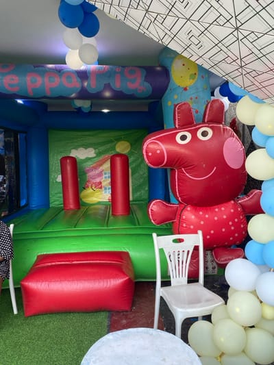The scene is an inflatable bounce house with a Peppa Pig theme. It is located in Bonadikombo, Cameroon, at coordinates (latitude: 4.048, longitude: 9.218). The bounce house is primarily blue and green with a red Peppa Pig character standing next to it. There are red inflatable poles in front of the bounce house. A white plastic chair is positioned in front of the bounce house, and several white, yellow, and blue balloons are placed in front of the chair. A red inflatable bench is located on the left side of the bounce house. An adult wearing a black and white polka dot shirt is partially visible sitting on the left side of the image. The bounce house is in an indoor setting with a white ceiling and some decorative white and black paper hanging from the ceiling.