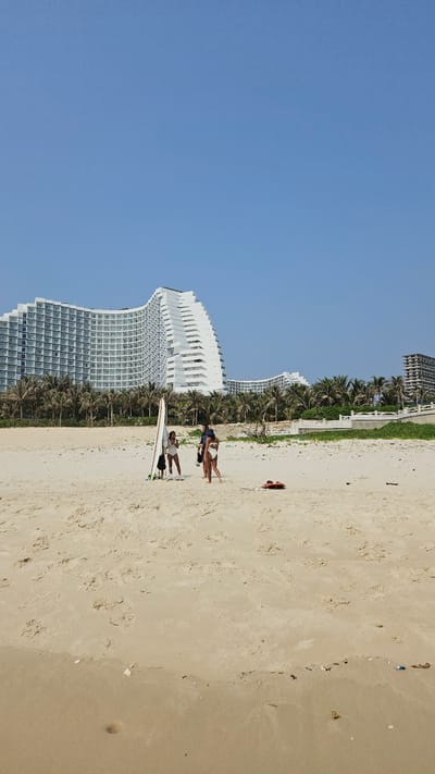 This image captures a vibrant, sunny day at a sandy beach in Bac Cam Ranh, Vietnam. The scene is dominated by a clear, bright blue sky and a wide expanse of light brown sand, stretching towards the right.

In the mid-ground, a series of modern high-rise buildings with numerous windows form the backdrop. A particularly striking white building exhibits a distinctive curved, wave-like architectural design. Further along, another tall, white structure, possibly under construction or renovation, is visible. These buildings are separated from the beach by a lush line of green palm trees and other dense foliage, suggesting a resort or urban coastal area.

On the sandy beach, four individuals are present. Closer to the foreground and the palm trees, two people dressed in swimwear are standing near a white surfboard, which is propped upright. One person appears to be interacting with the board or the other individual. Further down the beach, two more people are seen. One stands near a red object, possibly a rescue buoy or a small inflatable raft, while the other figure appears to be sitting or lying on the sand beside it.

The sand itself shows numerous footprints, indicating recent pedestrian activity. Small pieces of debris or shells are scattered across the beach. The clear sky and bright light suggest it is daytime with good weather, creating a relaxed and leisurely atmosphere typical of a beach vacation destination. There is no visible text in the image.