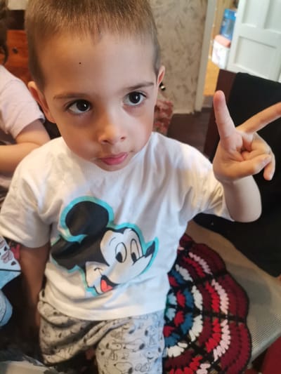 A young boy is posing indoors. He is wearing a white T-shirt with a Mickey Mouse graphic on the front, and gray patterned pants. He is holding his right hand up with his index and middle fingers extended, in a "peace" sign gesture. He is sitting on a surface covered with a multi-colored knitted blanket. The background features interior elements such as a door, parts of furniture, and a patterned wall. There is no specific information available to determine if the location is in the city of Pobeda, Bulgaria.