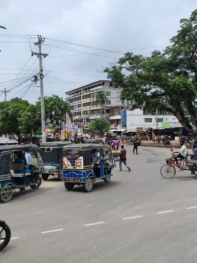 This is a street scene in Benapole, Bangladesh, featuring a busy intersection with multiple auto-rickshaws, a bicycle, and pedestrians. The road is paved with asphalt and marked with white lines. In the background, there are several multi-story buildings, some appearing to be commercial establishments, and lush green trees. Power lines and utility poles are visible overhead, with a dense network of wires. Several auto-rickshaws are parked or moving on the road, some carrying passengers. One auto-rickshaw in the foreground on the right is blue and yellow, with advertising on its side. A man is seen riding a bicycle with a red frame on the right side of the image. People are walking on the road and sidewalks. The sky is overcast, suggesting a cloudy day. The overall impression is one of a bustling urban environment in Bangladesh.