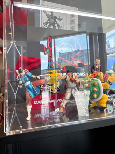 The image is an indoor shot of a display case exhibiting several action figures. In the foreground, a female action figure, possibly Chun-Li from Street Fighter, is depicted mid-kick, wearing a blue and white outfit with red leggings. To her right, a male action figure, resembling Ryu from Street Fighter, is in a fighting stance with red boxing gloves. Behind them, a Superman action figure stands with a red cape. Further back and to the right, a female character with blonde hair in a black and red outfit is visible, and a Bowser action figure from Super Mario is prominently displayed. The figures are arranged on a reflective surface, with a red display background featuring the text "TAMASHII NATIONS." Reflections of the surrounding environment, including windows and buildings, are visible on the glass of the display case. The overall setting appears to be a retail or exhibition space.