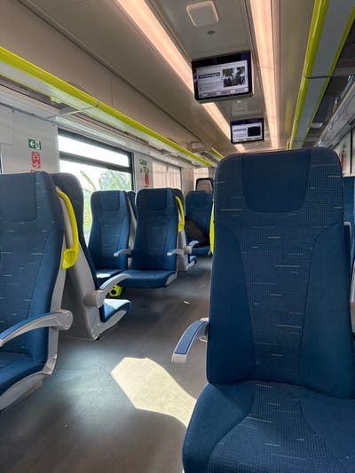The image shows the interior of a train car in Caravaggio, Italy, located at coordinates 45.483 latitude and 9.646 longitude. The train car is empty except for a single passenger seat in the foreground, with a blue and grey patterned fabric cover. The seat has a grey armrest on the left side and a yellow armrest on the right. The train car is lined with a series of five other seats, all with blue and grey fabric covers. The train car is well lit by overhead fluorescent lights. There are three windows on the left side of the train car. The window furthest back in the image is partially obscured by a thin green curtain. The window in the center of the image shows a glimpse of trees and buildings outside. The window at the front of the image is obscured by a yellow handrail. There are two overhead monitors above the train seats; one monitor displays a news report and the other monitor is displaying a blank screen.  The walls of the train car are painted white. The floor is covered in a light grey carpet. The train car is likely empty because it is not rush hour.  The train car is clean and well-maintained. There are no visible signs of wear or tear. There are no personal items visible in the train car. The train car appears to be relatively new.