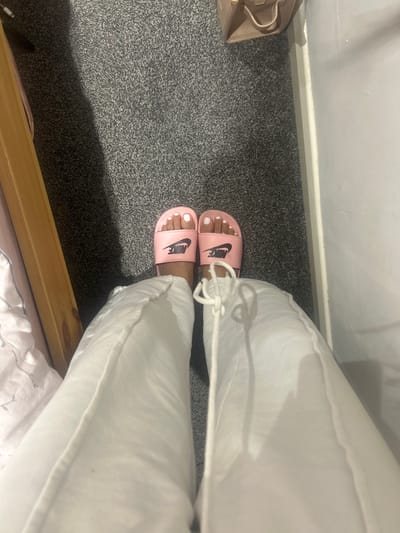 The image provides an overhead perspective of a person's lower legs and feet within an indoor environment, specified as Winslow, United Kingdom. The individual's feet are wearing light pink Nike slides, each bearing a black "NIKE" logo on the strap. The toenails on both feet are painted white. The person is dressed in white, loose-fitting trousers, with a visible white drawstring or tie detail on the front of the right leg. The floor is covered with a grey, short-pile carpet. To the left of the frame, a segment of light-colored wooden furniture, likely part of a bed frame, is partially visible next to white patterned bedding. To the right, a plain white wall extends vertically. In the upper right corner, a light-brown handbag or purse is resting on the carpet, leaning against the white wall.