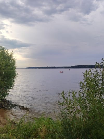 The image depicts a wide expanse of water, likely a reservoir, under a cloudy sky. In the foreground, lush green grass and foliage line the bank. To the left, a large, leafy tree with a visible trunk is positioned near the water's edge, where rocks and sand are present. The water itself is calm, with subtle ripples. In the middle distance, on the water, two people are engaged in water activities. One appears to be on a red paddleboard or kayak, and another individual is standing on a paddleboard further out. The far bank of the water is lined with a dense forest. The overall scene is natural and serene, with a sense of calm before potential weather changes indicated by the heavy clouds. The location is identified as the Klyazma Reservoir Tourist Boarding House, Russia.