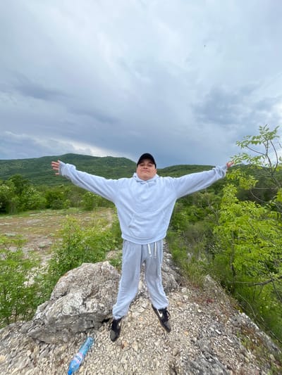A young man stands on a rocky precipice, arms outstretched in a pose of exuberance or balance. He is dressed in a light grey hoodie and sweatpants, with a black baseball cap and black sneakers. The backdrop consists of lush, green hills and a cloudy sky. The foreground includes loose rocks and greenery. A partially visible plastic water bottle sits near his feet. The location is Kamchia, Bulgaria, as indicated by the brand on the water bottle "Devin" which is a bottled water company operating in the area.