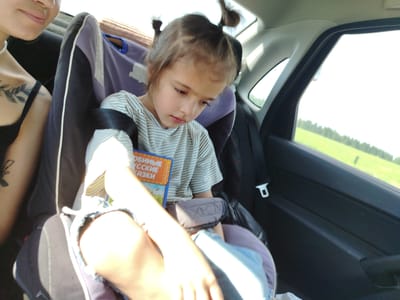 In a vehicle traveling on a road, a young girl with two high pigtails sits in a car seat. She is wearing a striped t-shirt and denim shorts, and she holds a book with a colorful cover that reads "Любимые русские сказки" (Beloved Russian Fairy Tales). To her left, a woman with a nose ring and a tattoo on her shoulder is partially visible. The background shows a bright sky and a green, tree-lined landscape, suggesting the car is in a rural area near Malyi Gondyr, Russia. The image captures a casual moment of travel, with the child seemingly engrossed in her book.