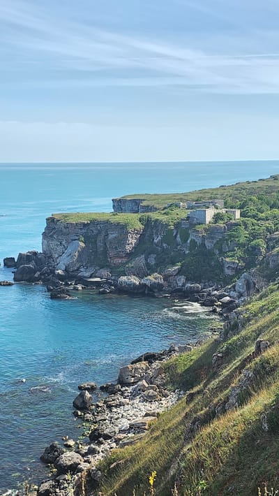 The image shows a scenic view of the Kamen Bryag coast in Bulgaria. Cliffs are visible to the right of a tranquil ocean landscape. An old building complex rests on the higher cliffs. The landscape is composed of grassy areas and rocky terrain.