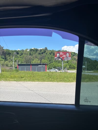 The image presents a bright, sunny view from inside a vehicle, looking out onto a roadside scene in Abgharkhuk, Abkhazia. The foreground shows parts of the car's dark interior and a blue-tinted window, framing the landscape.
An asphalt road runs along the right side of the frame, alongside a verdant grassy shoulder. Beyond the road, dense green trees and foliage cover rolling hills, suggesting a natural, possibly mountainous, environment under a clear blue sky dotted with fluffy white clouds.
A prominent billboard in the midground displays two portraits of men against a red and orange patterned background. The top portrait is labeled "АХБА ДАВИД АНДРЕЕВИЧ" (AKHBA DAVID ANDREEVICH), and the bottom one is "АХБА САНДР СИТОВИЧ" (AKHBA SANDR SITOVICH), depicted wearing a uniform with visible medals.
Further along the road, a dark-colored building with red vertical stripe accents is visible. A white sign on its side bears the Abkhazian text "ШХӘЫОНЧХӘ" (SHKHYONCHKHV). A white car is parked near the billboard. Utility poles and wires are also interspersed throughout the scene. The time of day appears to be midday, characterized by strong sunlight and clear weather.