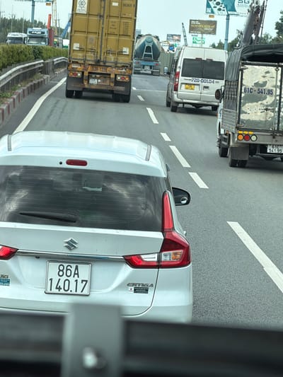 This media file depicts a street-level view of traffic in Dong Nai City, Vietnam. The primary subjects are various vehicles on a paved road, suggesting a daytime setting. In the foreground, the rear of a white Suzuki Ertiga is visible, displaying a license plate "86A 140.17". To its left, a yellow shipping container truck is prominent, with license plate "53R-1226" and additional markings like "563921 8" and "PAY LOAD". Further in the background, a white van with license plate "72G-001.04" is observed. The road has white lane markings, and a concrete barrier with red and white reflectors separates the lanes. Beyond the road, some greenery and buildings, including what appears to be construction equipment and signs with Vietnamese text like "HOA VIÊN BÌNH AN", are visible. The scene conveys a typical urban traffic environment. The image orientation is landscape, with the main focus on the vehicles and the road. No people are clearly identifiable in the scene. The weather appears to be clear and the time of day is likely daytime, given the lighting conditions.
