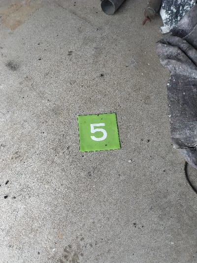 The scene captures a downward view of a textured, light gray concrete floor. A square, bright green marker, approximately 10-15 cm per side, featuring a prominent white numeral '5', rests flat in the center-lower portion of the frame. The concrete exhibits varying shades of gray, consistent with wear and minor soiling, including scattered dark granular debris and faint stains. In the upper right corner, a dark gray cylindrical pipe is partially visible, alongside a smaller, rusted metallic object with a pointed tip. Adjacent to these, a crumpled mass of dark gray fabric, possibly a heavy-duty cloth or tarpaulin, shows white speckled residue. No individuals are present. The environment suggests an indoor or sheltered utility area, such as a garage or workshop. No specific landmarks or unique features are present to precisely identify the location within Bitburg, Germany.