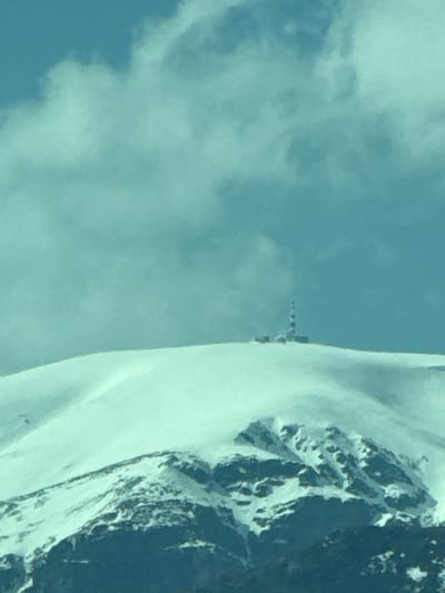 The image shows a snowy mountain ridge under a cloudy sky. A line of small objects, potentially a path or trail, can be seen leading up the ridge. The location of Manolovo, Bulgaria is not evident from the image.