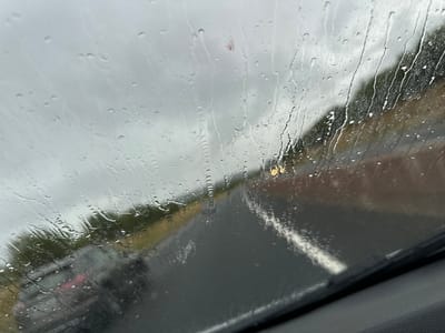 The image shows a view through a rain-streaked car windshield, looking out onto a wet road. On the left, a dark-colored vehicle is visible, its taillights illuminated. Further ahead, traffic can be seen on the road, with a single yellow traffic light visible in the distance. The sky is overcast and gray. The car appears to be on a road with trees lining the side, suggesting a location in a somewhat rural or suburban area, potentially near Clonmore Court, Ireland, given the context of the query. The overall impression is one of traveling in rainy weather.