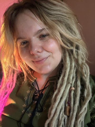 This media file shows a close-up, angled shot of a person with blonde hair and dreadlocks. The person's face is partially obscured by their hair, but they are looking towards the camera with a slight smile. They are wearing a green garment with embroidered details. The lighting in the image is warm, with pink and orange hues reflecting on the person's hair and skin, suggesting an artificial light source or a colorful environment. The overall mood appears relaxed and intimate. There is no visible text in the image. The setting appears to be indoors, possibly a room with colorful lighting.