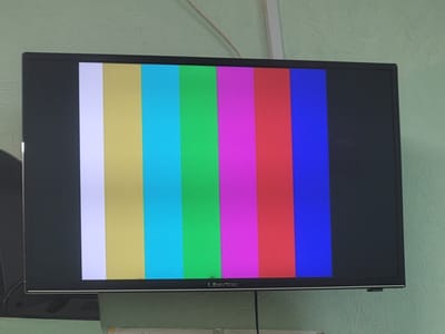 The image shows a television screen displaying a color bar test pattern. The screen is black with vertical stripes of white, yellow, cyan, green, magenta, red, and blue. The television set is mounted on a wall painted a light green color. The brand name "Liberton" is visible on the bottom bezel of the television. The scene appears to be indoors. There are no people or animals visible in the image. The image was taken in Khotsky, Ukraine.